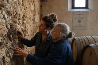 Two women in a Provence winery examining an exposed soil wall, softly lit, deeply engaged in discussion; calm, authentic atmosphere.