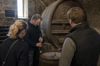 Authentic moment in a Champagne cellar as visitors quietly watch a vintner uncork a bottle near old barrels, afternoon light softly filtering in.