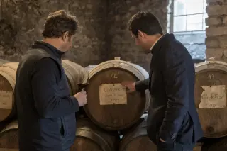Sommelier and vigneron discussing over old barrels in a softly lit stone cellar, natural light filtering in, authentic and contemplative atmosphere.
