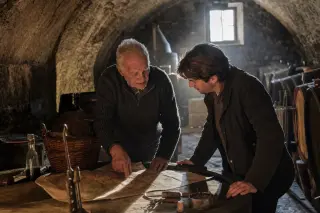 In a stone wine cellar lit by natural morning light, an older winemaker and a guest study a vineyard map on a barrel, surrounded by winemaking tools, sharing a quiet moment of discovery.