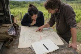 Two people consult a detailed wine region map on a vehicle tailgate beside a vineyard, planning their wine journey under soft natural light.